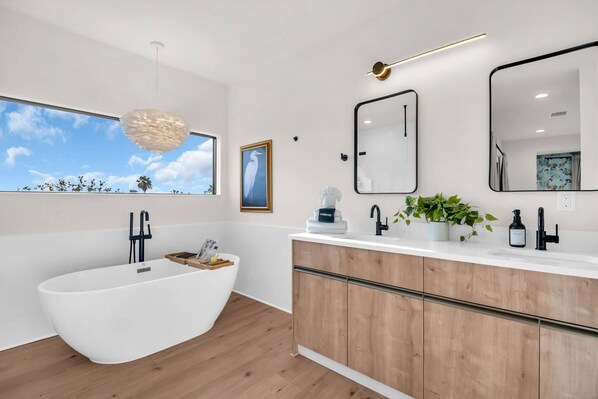 Ein modernes Badezimmer mit einer freistehenden Badewanne, Holzschränken, einem großen Spiegel und Blick auf Palmen.