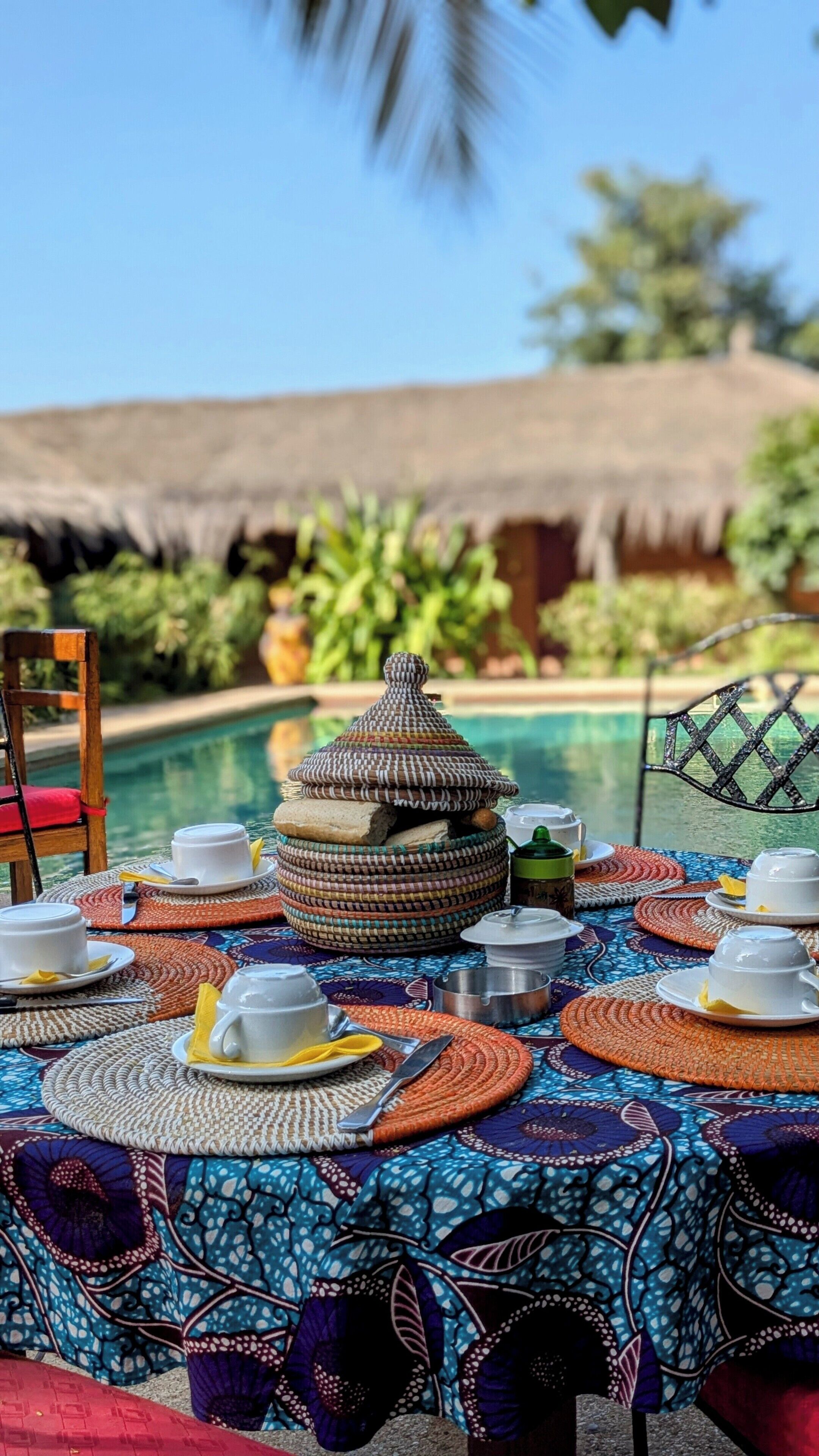 An outdoor dining area with a pool, a thatched-roof structure, and a table set with plates, cups, and a decorative container.