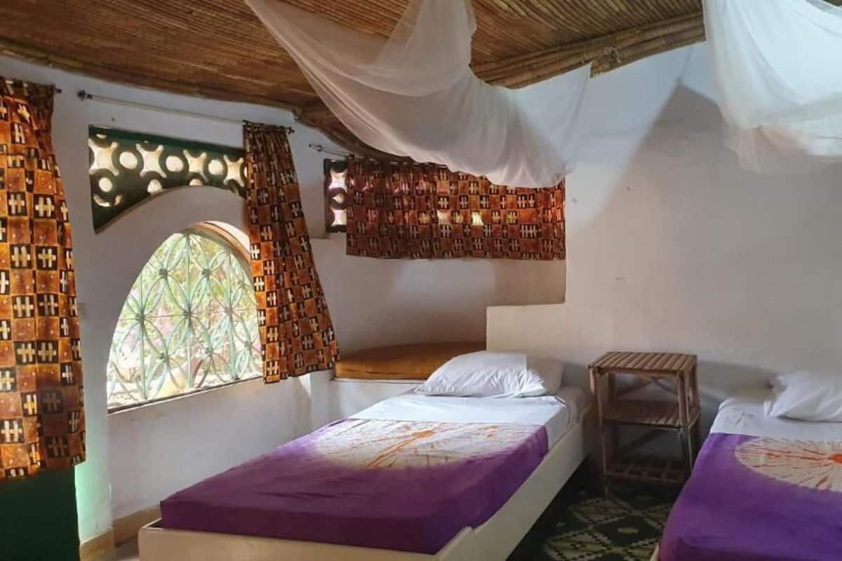 Quarto com duas camas, uma mesinha e janela com cortinas estampadas.