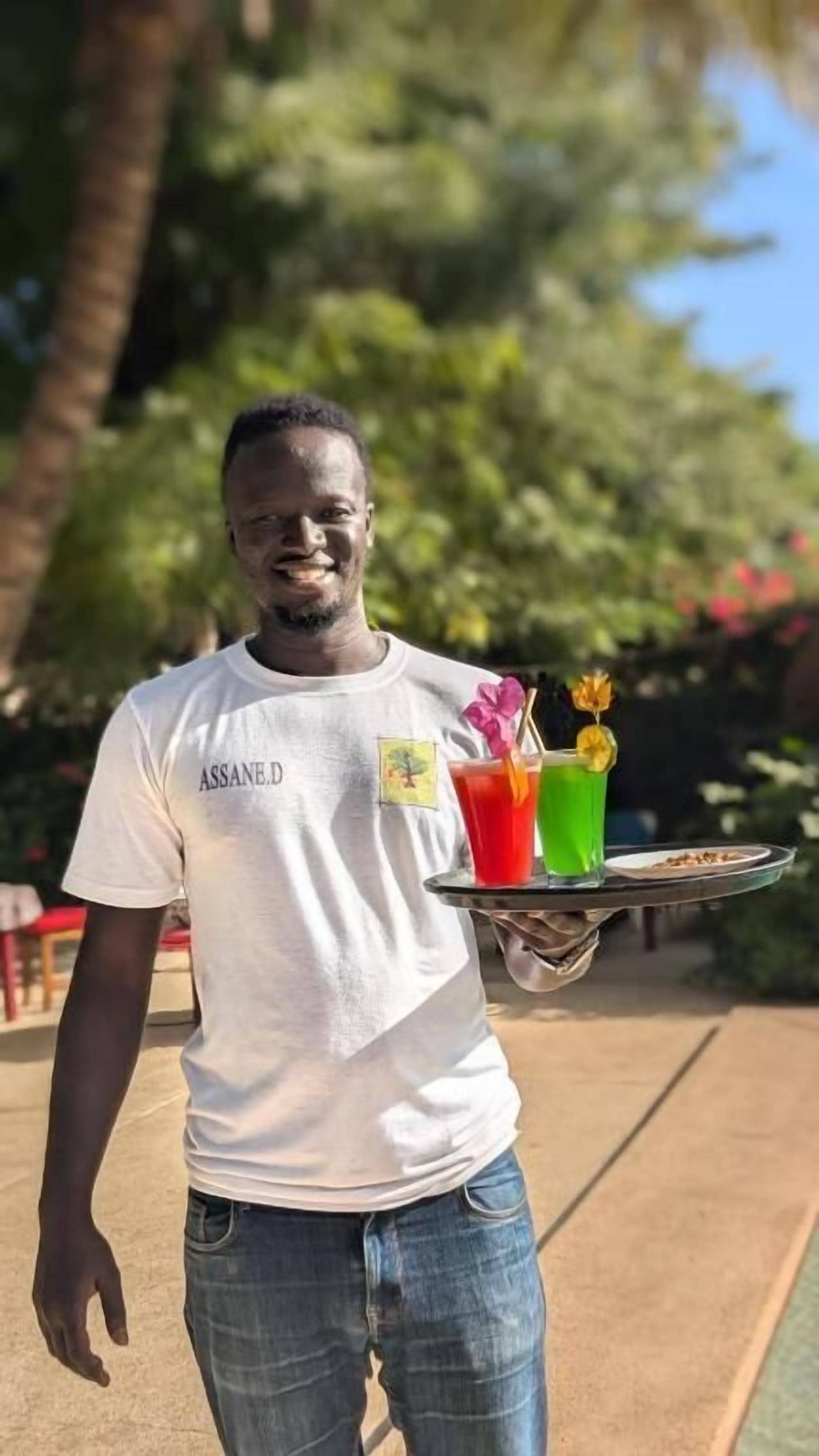 A person holding a tray with colorful drinks and a dessert, wearing a white t-shirt with text and a graphic, standing outdoors with greenery in the background.