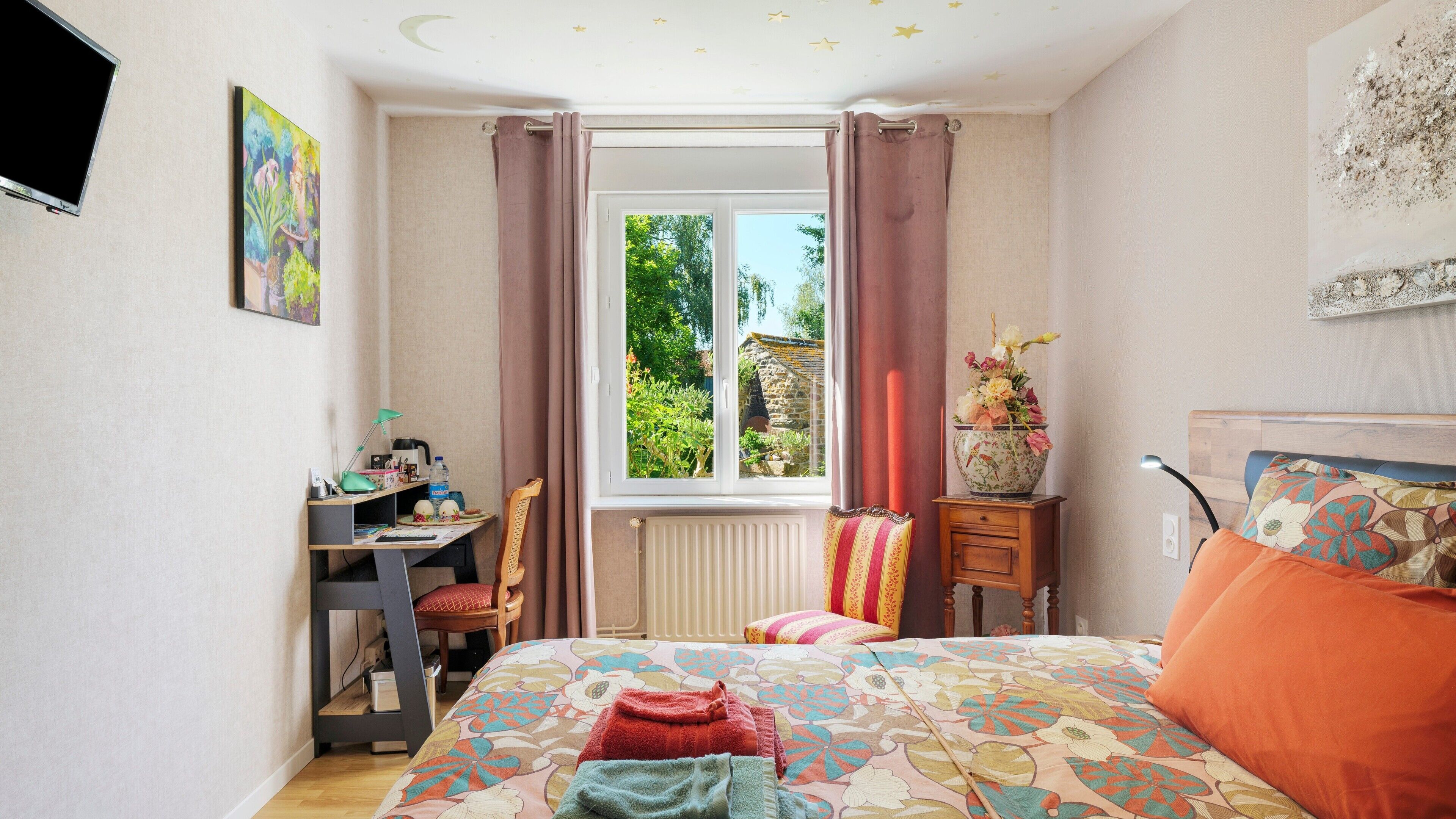 Une chambre à coucher comprenant un lit, un bureau, une chaise, un radiateur, une fenêtre avec des rideaux et un vase avec des fleurs.