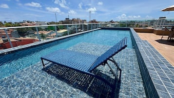 A rooftop swimming pool with a glass barrier, a lounge chair, and a view of the cityscape.