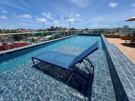A rooftop swimming pool with a glass barrier, a lounge chair, and a view of the cityscape.