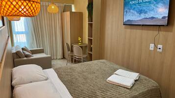 Quarto de hotel com cama, televisão exibindo 'PORTO DE GALINHAS', sofá, poltrona, mesinha com uma planta e guarda-roupa de madeira.