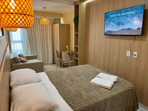 A hotel room with a bed, a TV displaying 'PORTO DE GALINHAS,' a sofa, a chair, a small table with a plant, and a wooden wardrobe. - Waikiki Garden (Ipojuca)