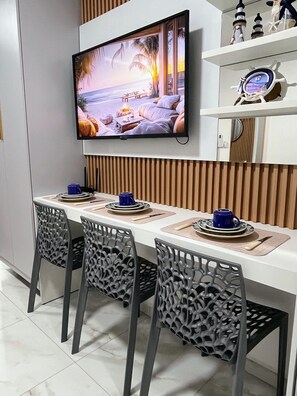 A modern dining area with a flat-screen TV mounted on the wall, a wooden panel wall, and a dining table set with chairs and plates. - Waikiki Garden (Ipojuca)