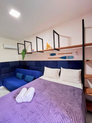 Ein Zimmer mit einem Bett, einem blauen Sofa, einer violetten Decke und einem an der Wand befestigten Regal mit diversen Gegenständen.