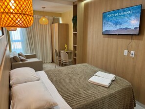 A hotel room with a bed, a TV displaying 'PORTO DE GALINHAS,' a sofa, a chair, a small table with a plant, and a wooden wardrobe. - Waikiki Garden (Ipojuca)