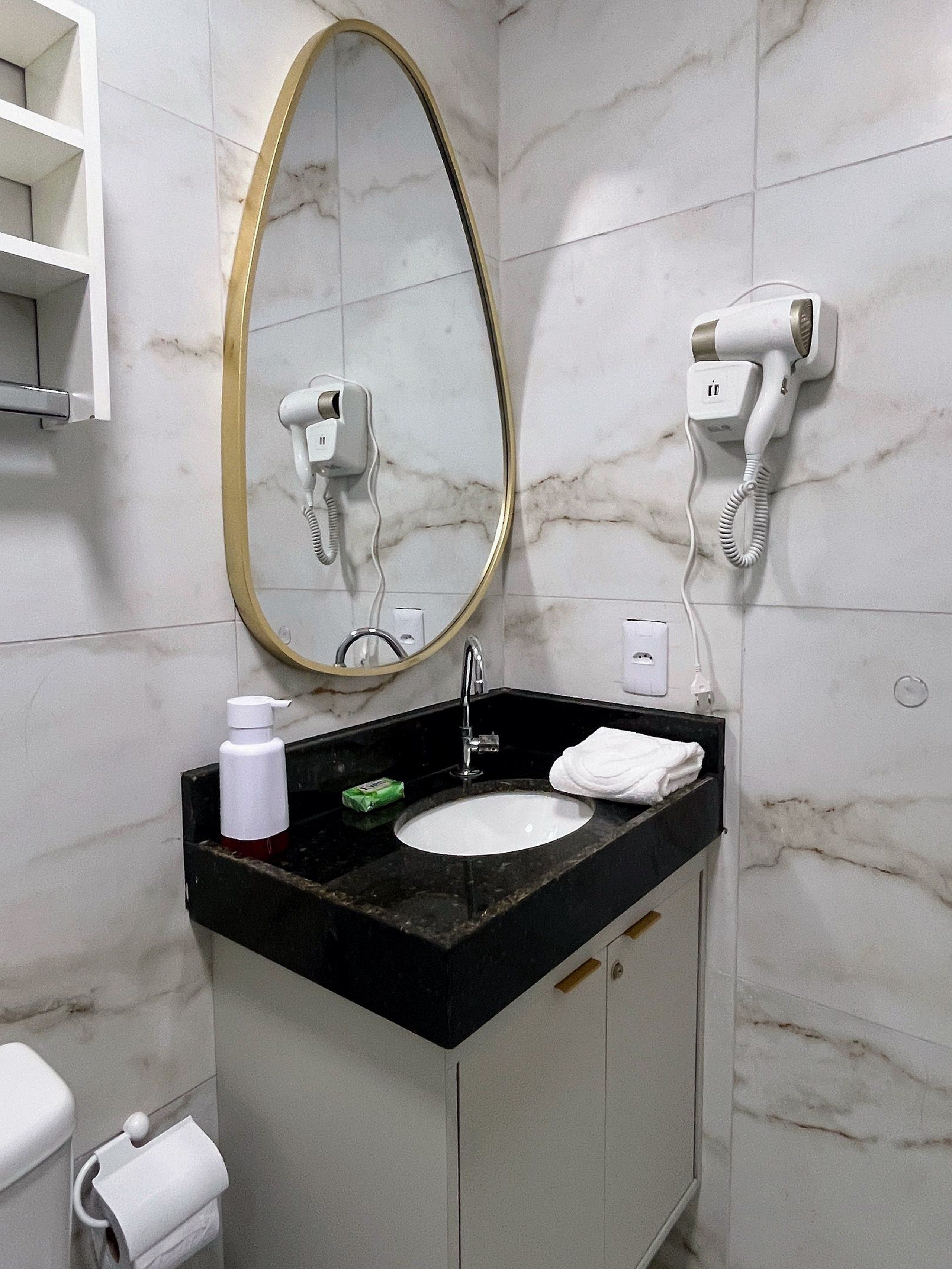 Une salle de bain avec un mur en marbre, un lavabo à vasque avec un plan de travail noir, un miroir avec un cadre doré et deux sèche-cheveux fixés au mur.