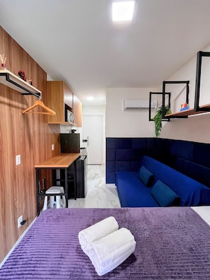 A compact room with a bed, a small kitchenette, a blue sofa, and a wooden wall. - Waikiki Garden (Ipojuca)