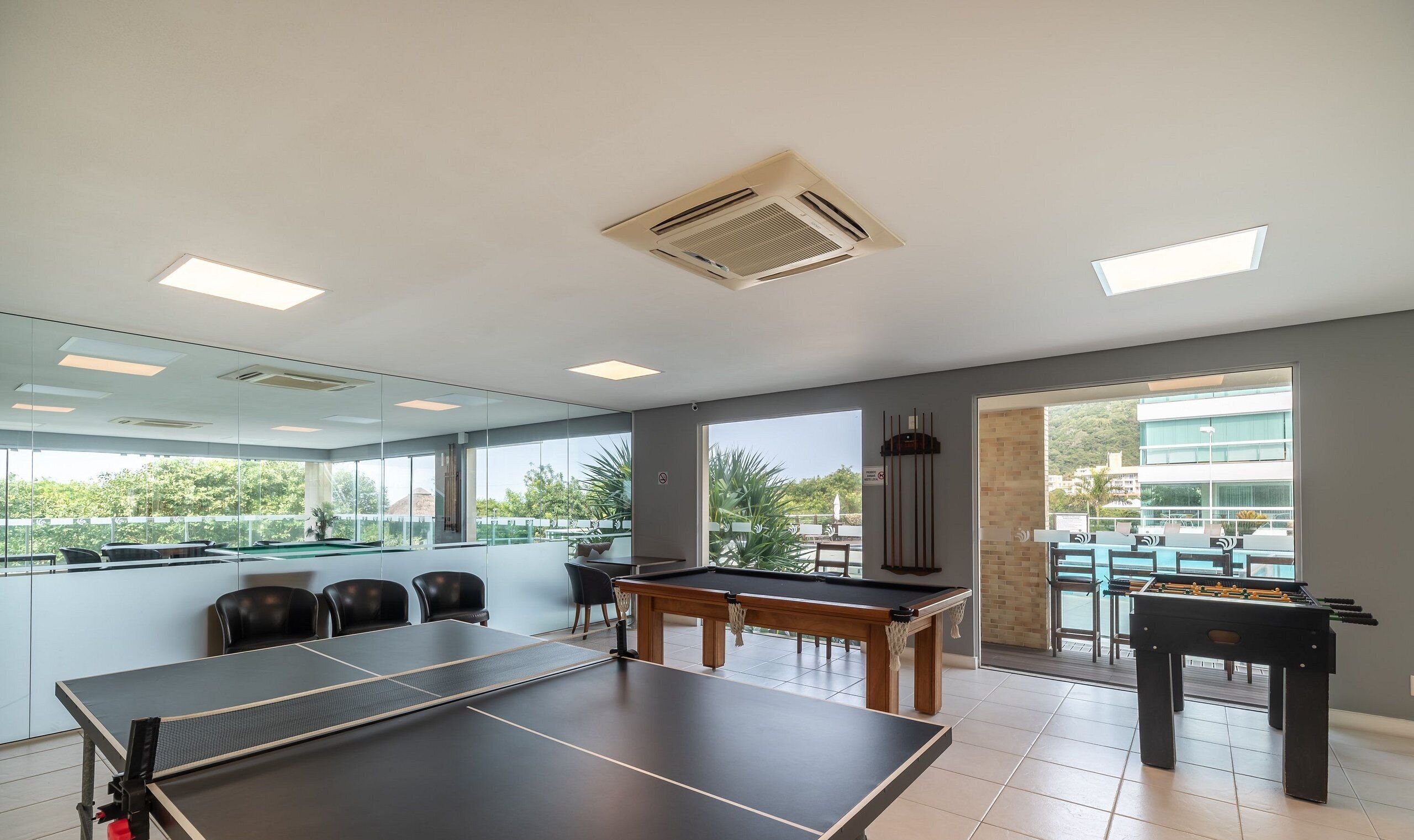 A spacious indoor area with a ping pong table, a pool table, and a foosball table, featuring large windows and a view of a pool and greenery outside.