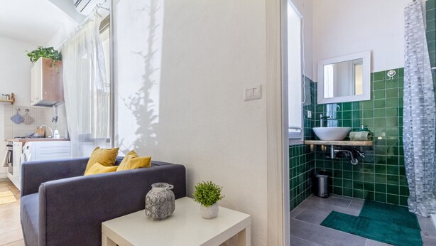 Eine moderne Wohnung mit Wohnbereich, ausgestattet mit einer Couch und einem kleinen Tisch, einer Küche mit grünen Fliesen und einem Badezimmer mit Duschvorhang.
