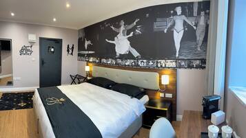 A hotel room with a bed, a desk, a chair, a coffee machine, and a wall with dance-themed artwork.