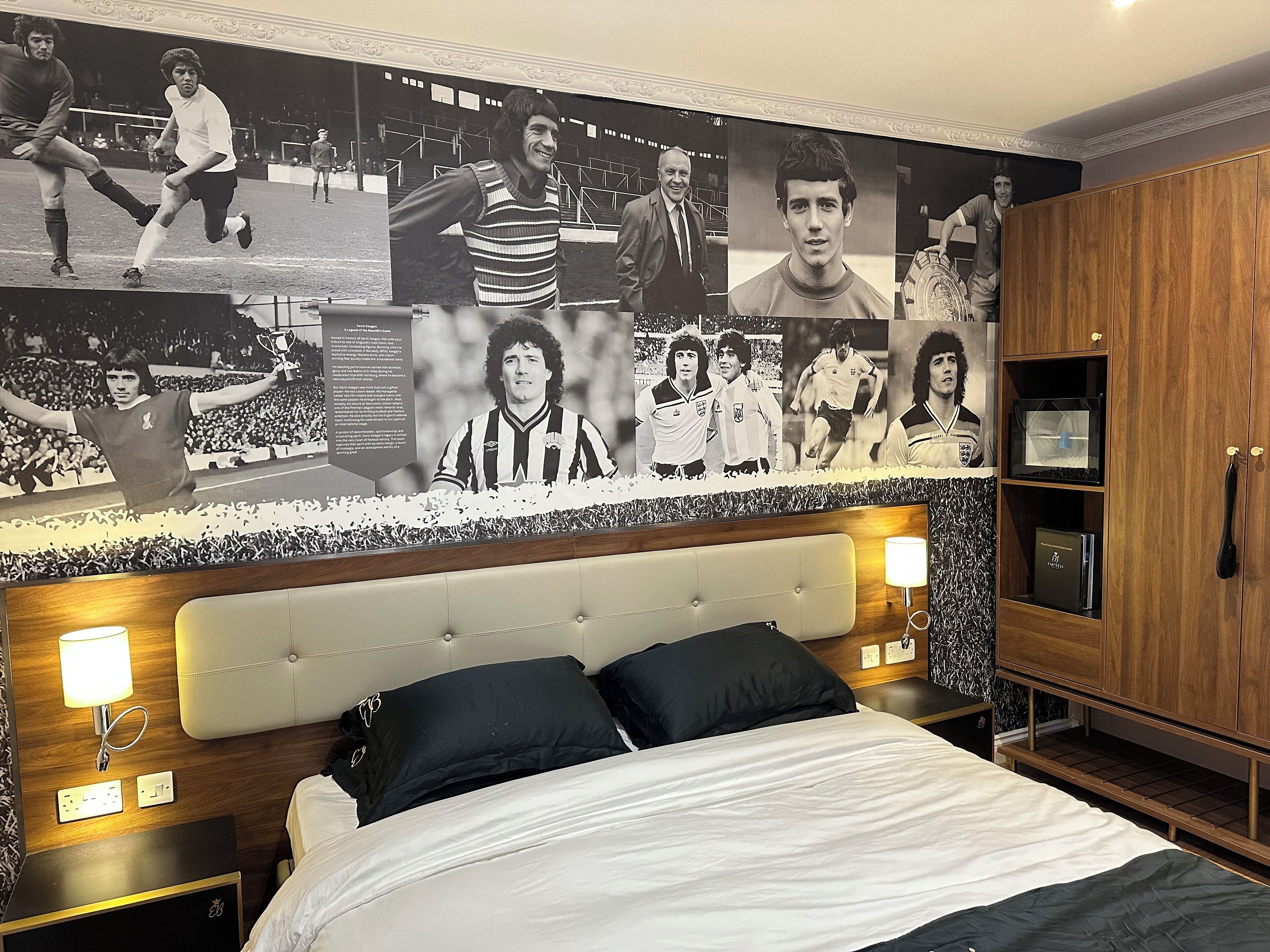 Ein Hotelzimmer mit einer großen Wandmalerei von Fußballspielern, einem gesteppten Kopfteil, einem Bett mit schwarz-weißer Bettwäsche und einem Holzschrank.