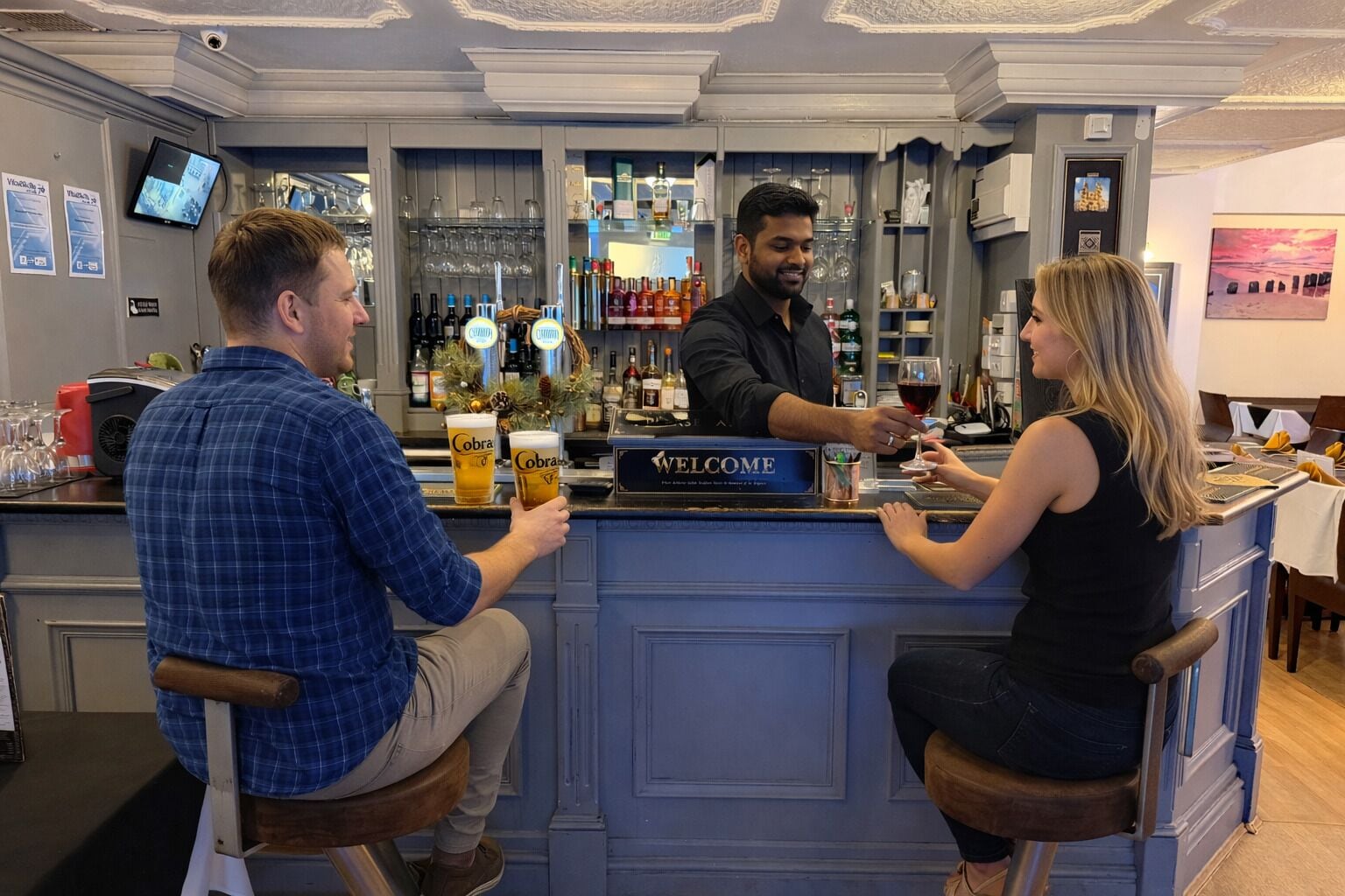 Uma área de bar com um letreiro 'WELCOME', banquetas e um balcão com um bartender.