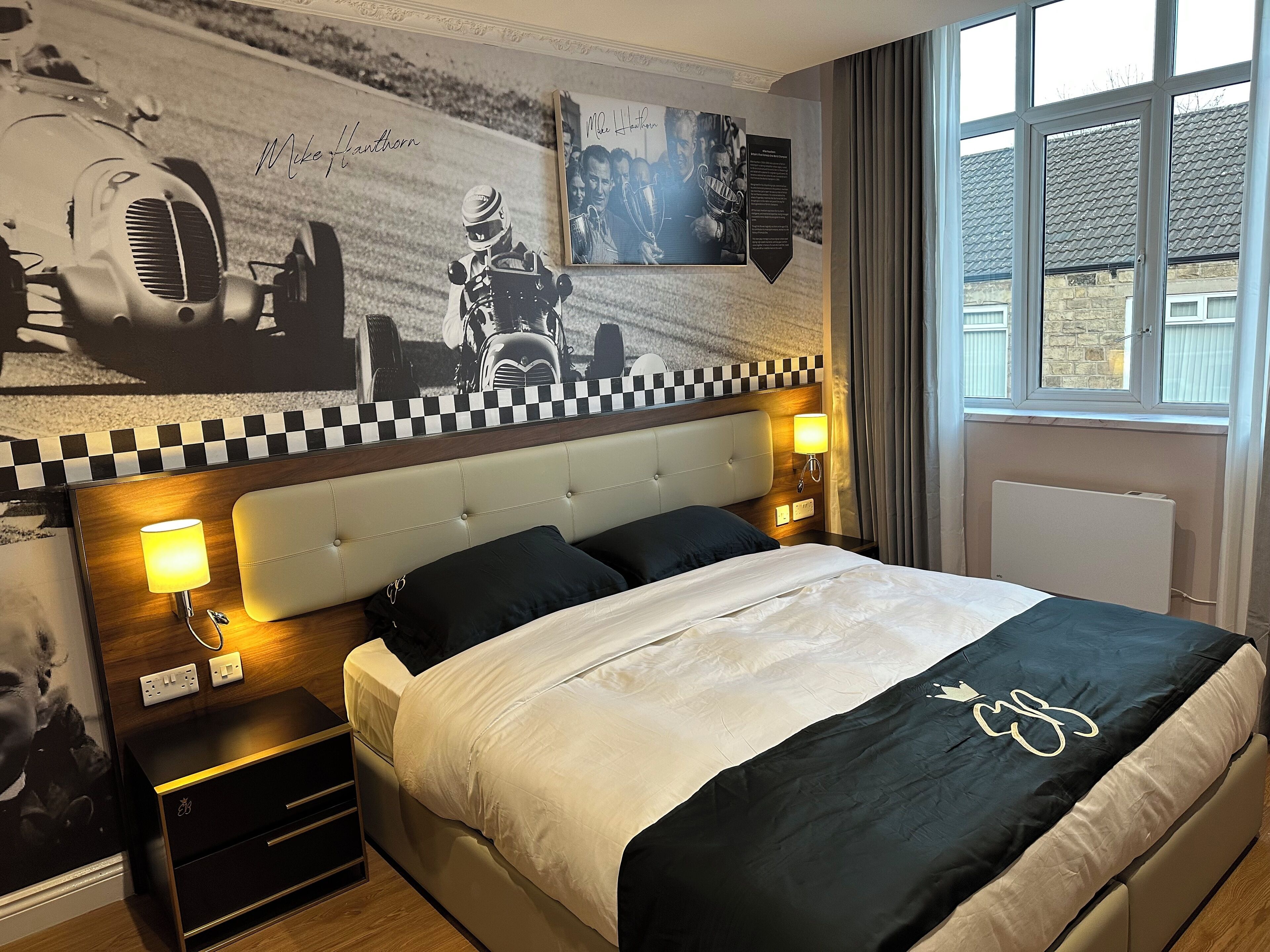 Una habitación de hotel con un cabecero a cuadros, una cama con colcha blanco y negro, una mesita de noche con lámpara y una pared con recuerdos de carreras.