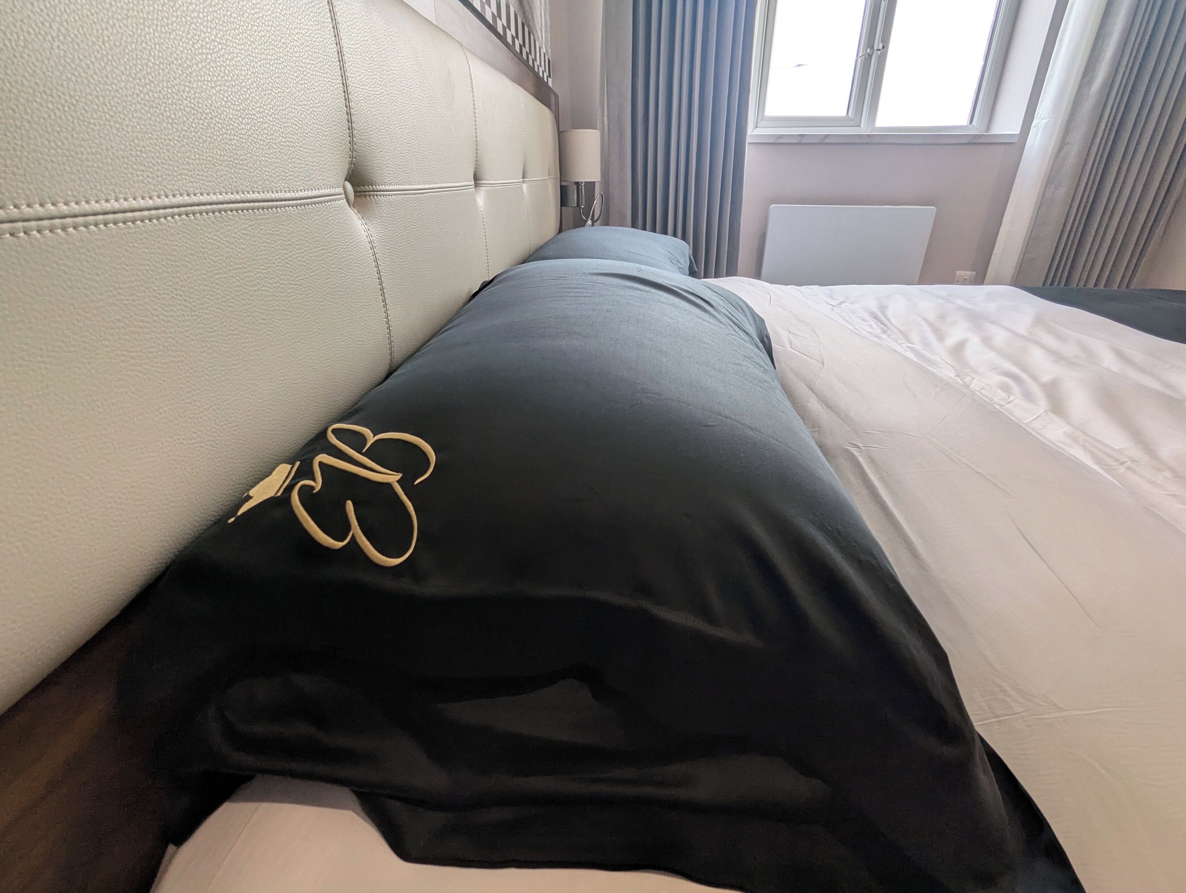 A hotel room with a tufted headboard, a black pillow with a gold embroidered design, and a white bedspread.