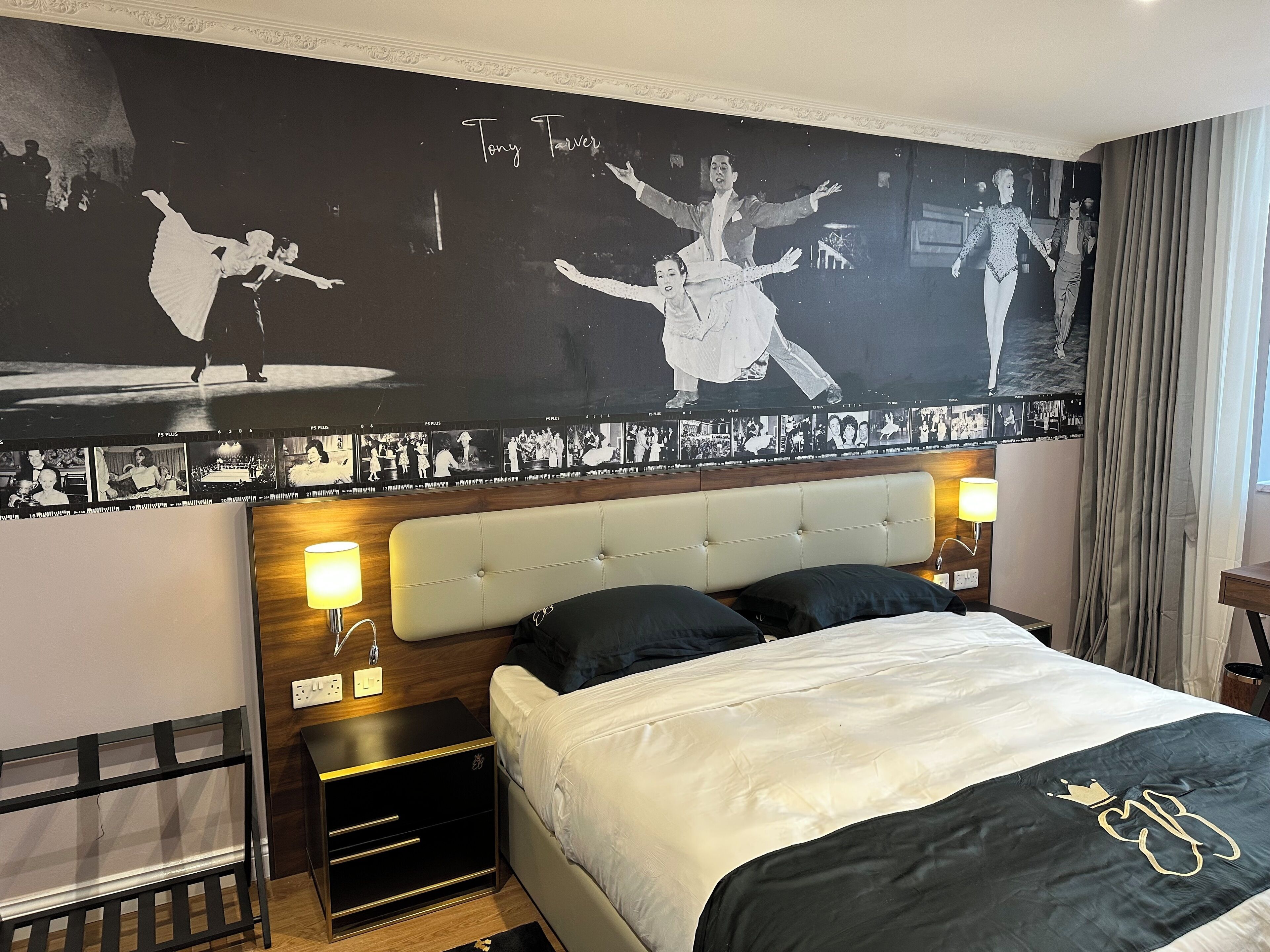 Habitación de hotel con un mural fotográfico grande en blanco y negro, un cabecero capitonado, dos lámparas de noche, una cama con colcha blanco y negro, y una mesita de noche con lámpara.