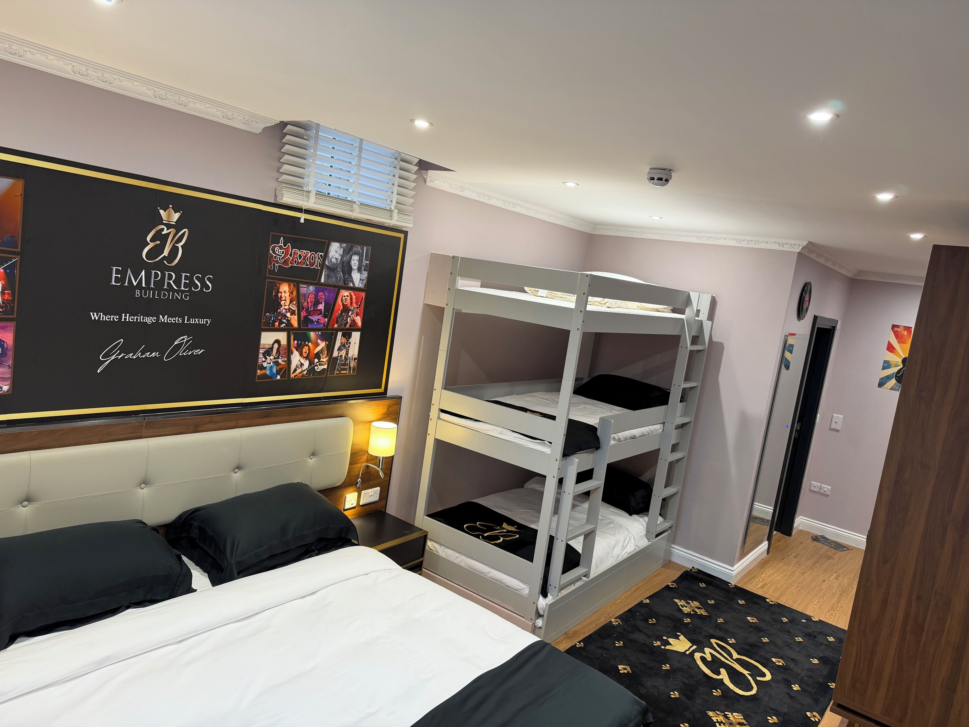 A hotel room with a bunk bed, a bed with black and white bedding, a headboard, a wall with a framed photo collage, and a small rug with a logo.