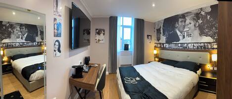 A hotel room with two beds, a desk, a flat-screen TV, and large framed pictures on the walls.