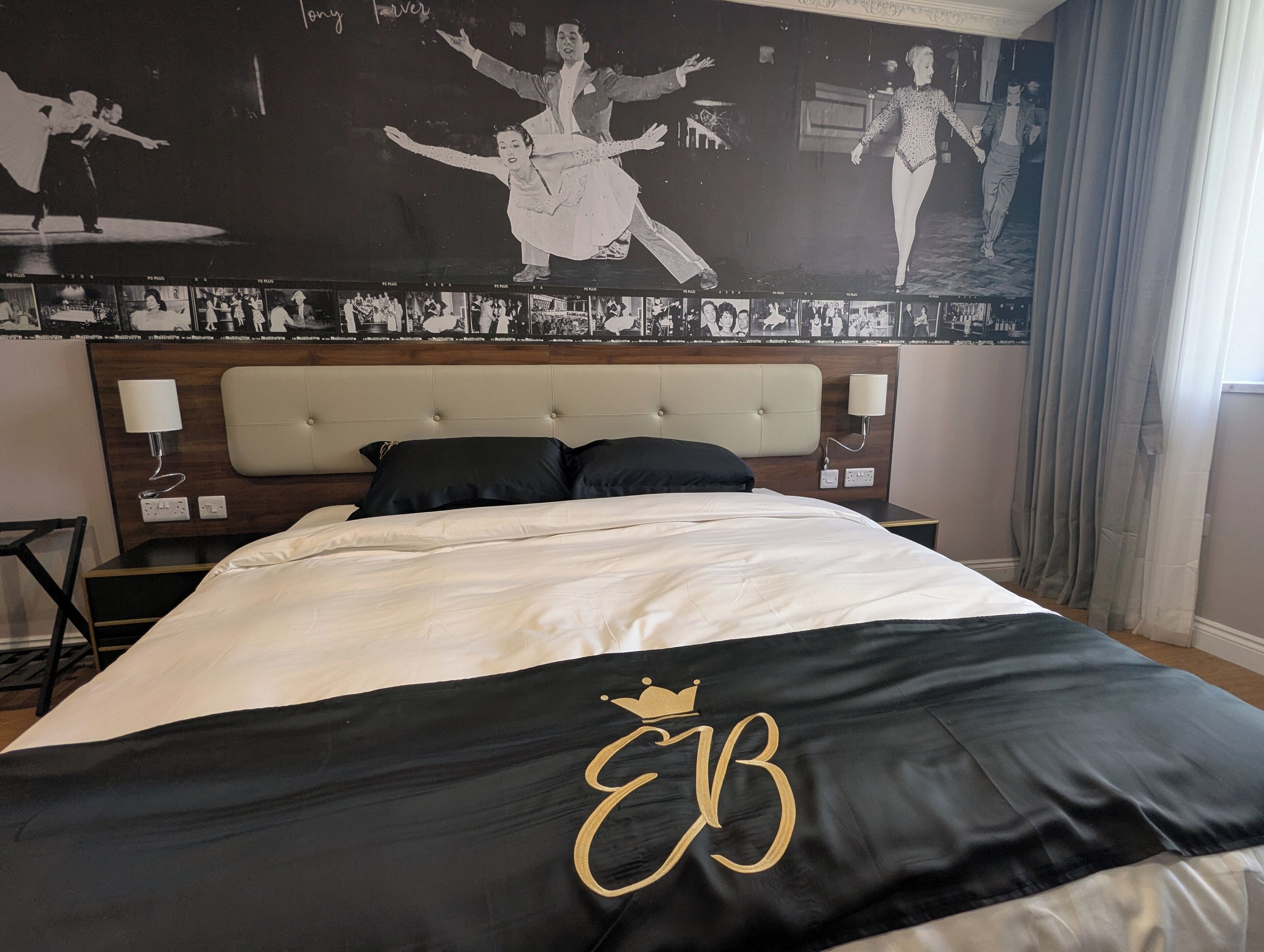 Habitación de hotel con una cama grande, cabecera con panel de color claro, dos mesitas de noche con lámparas y una pared con un collage de fotos en blanco y negro.