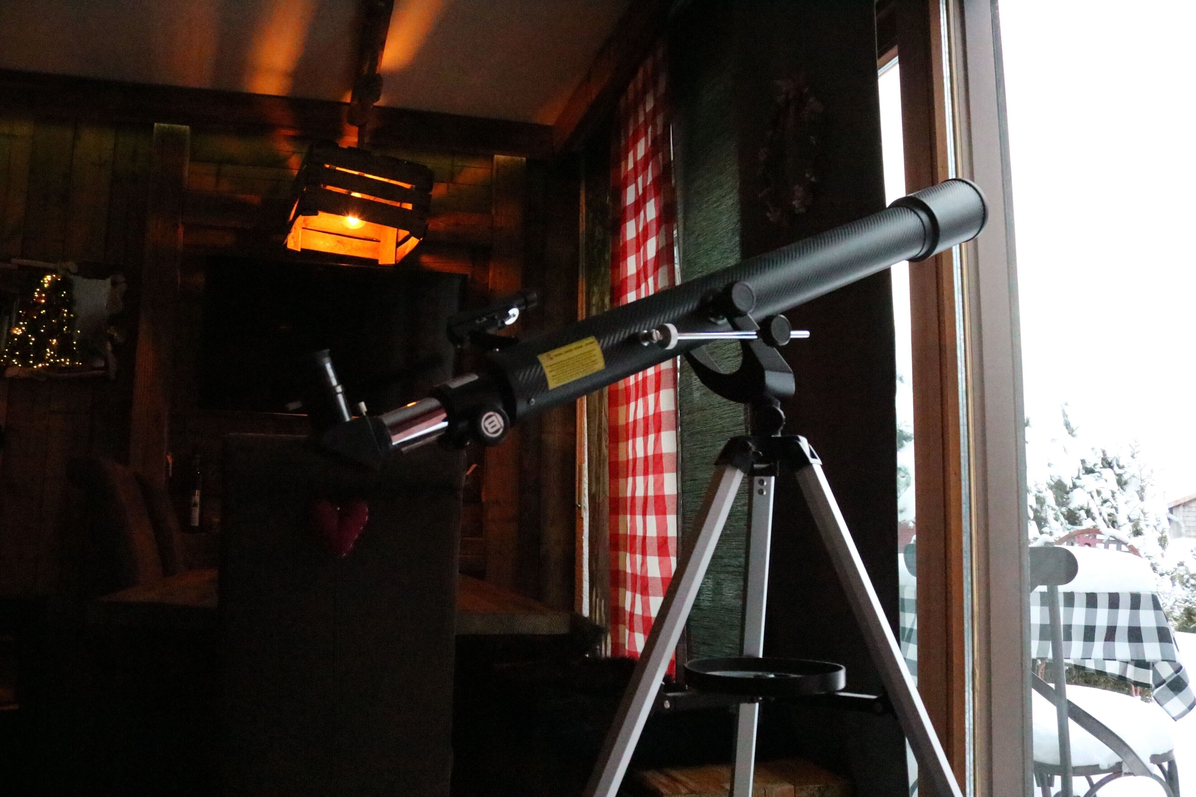 A telescope on a tripod in a room with a window, a checkered tablecloth, and a Christmas tree.
