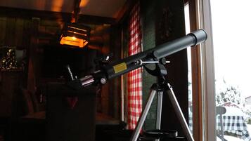 A telescope on a tripod in a room with a window, a checkered tablecloth, and a Christmas tree.