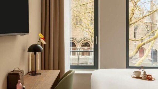 A hotel room with a bed, a desk, a chair, a TV, and large windows with a view of buildings and trees. - Hotel Middelland Rotterdam (Rotterdam)