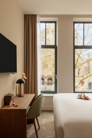 A hotel room with a bed, a desk, a chair, a TV, and large windows with a view of buildings and trees. - Hotel Middelland Rotterdam (Rotterdam)