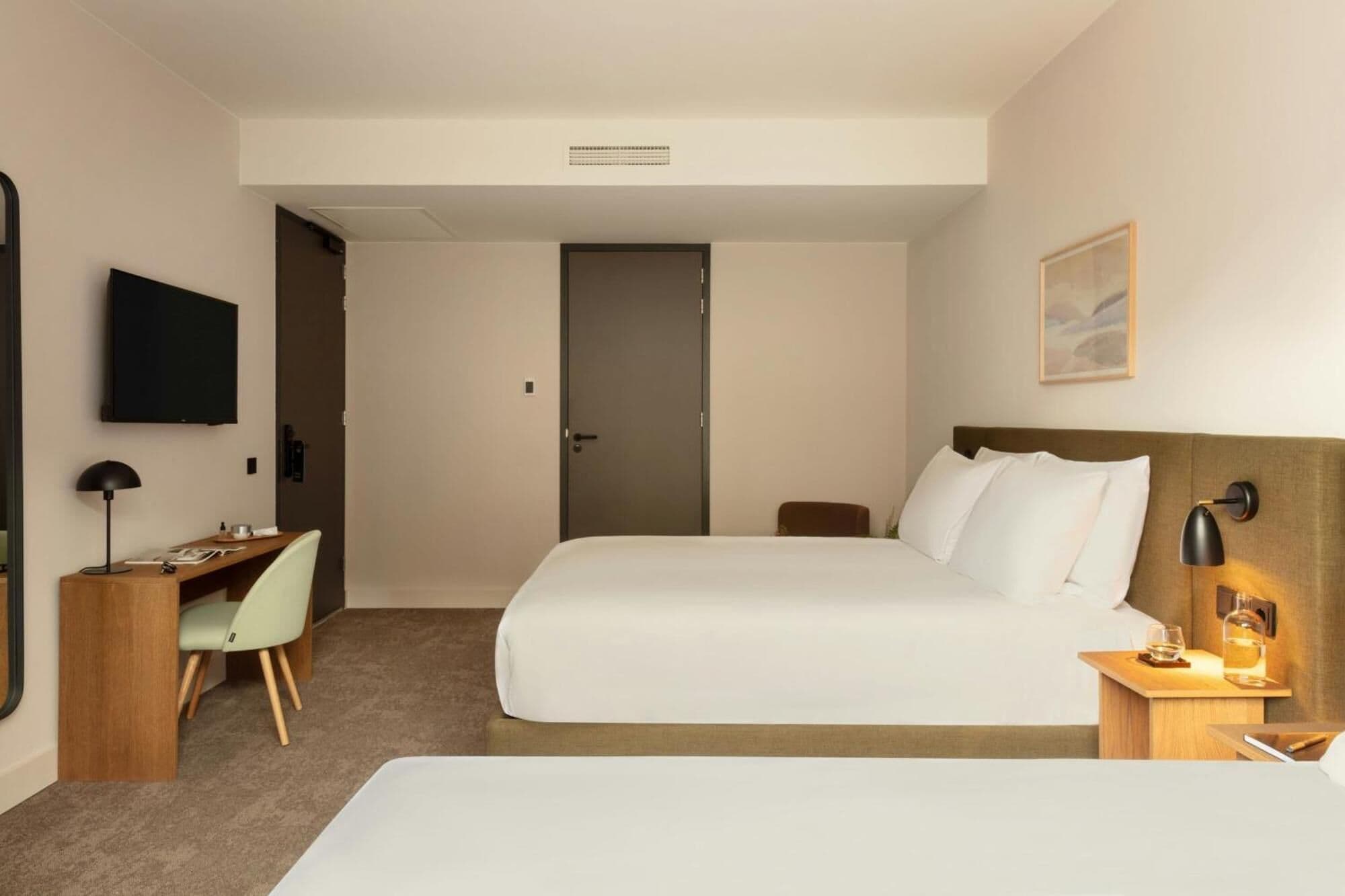 Quarto de hotel com uma cama, uma escrivaninha com cadeira, uma televisão e uma mesinha de cabeceira com abajur.