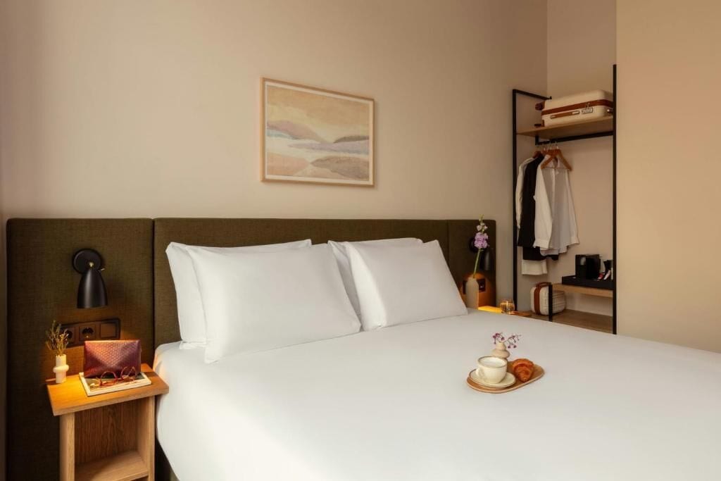 A neatly made bed with white linens, a bedside table with a lamp, a small vase with flowers, and a tray with a teapot and cup.