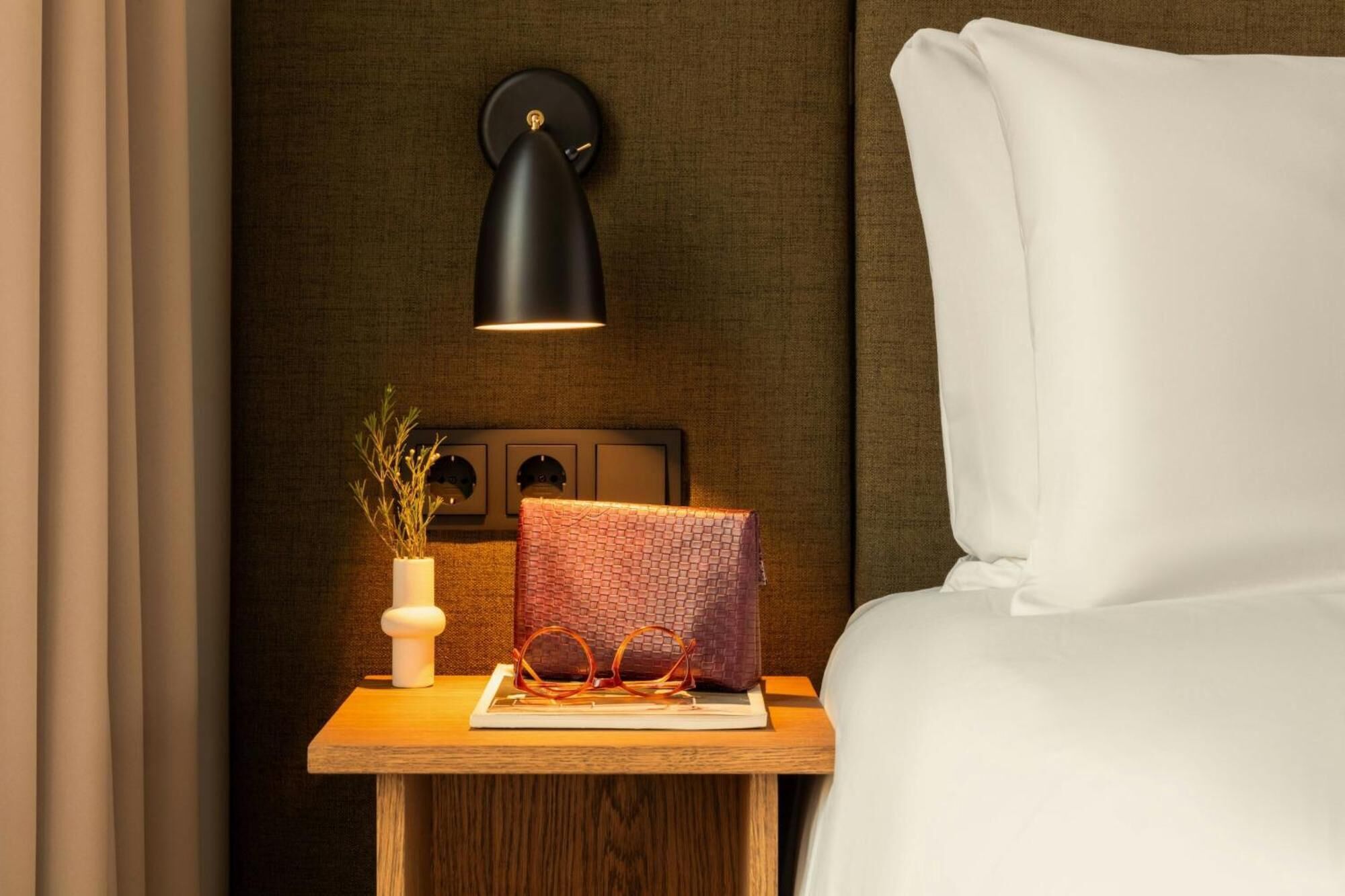 Cama bem arrumada com lençóis brancos, uma pequena mesa de cabeceira de madeira com uma luminária preta fixada na parede, um vaso branco com plantas secas e um objeto vermelho com textura na mesa de cabeceira.