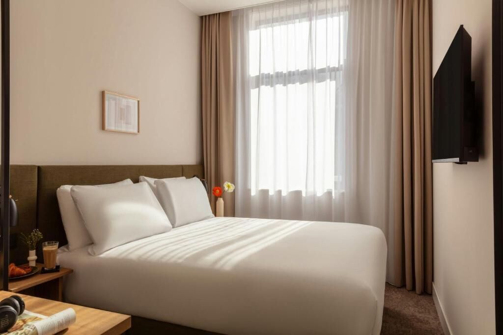 Una cama bien hecha con sábanas blancas en una habitación de hotel, una mesita de noche con una lámpara y un libro, y un ventanal con cortinas translúcidas.