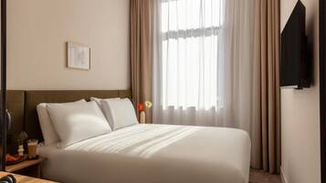 Una cama bien hecha con sábanas blancas en una habitación de hotel, una mesita de noche con una lámpara y un libro, y un ventanal con cortinas translúcidas.