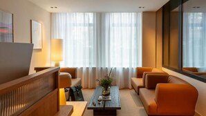 A modern hotel room with a wooden reception desk, a sofa set, a coffee table with a vase and a book, and large windows with sheer curtains. - Hotel Middelland Rotterdam (Rotterdam)