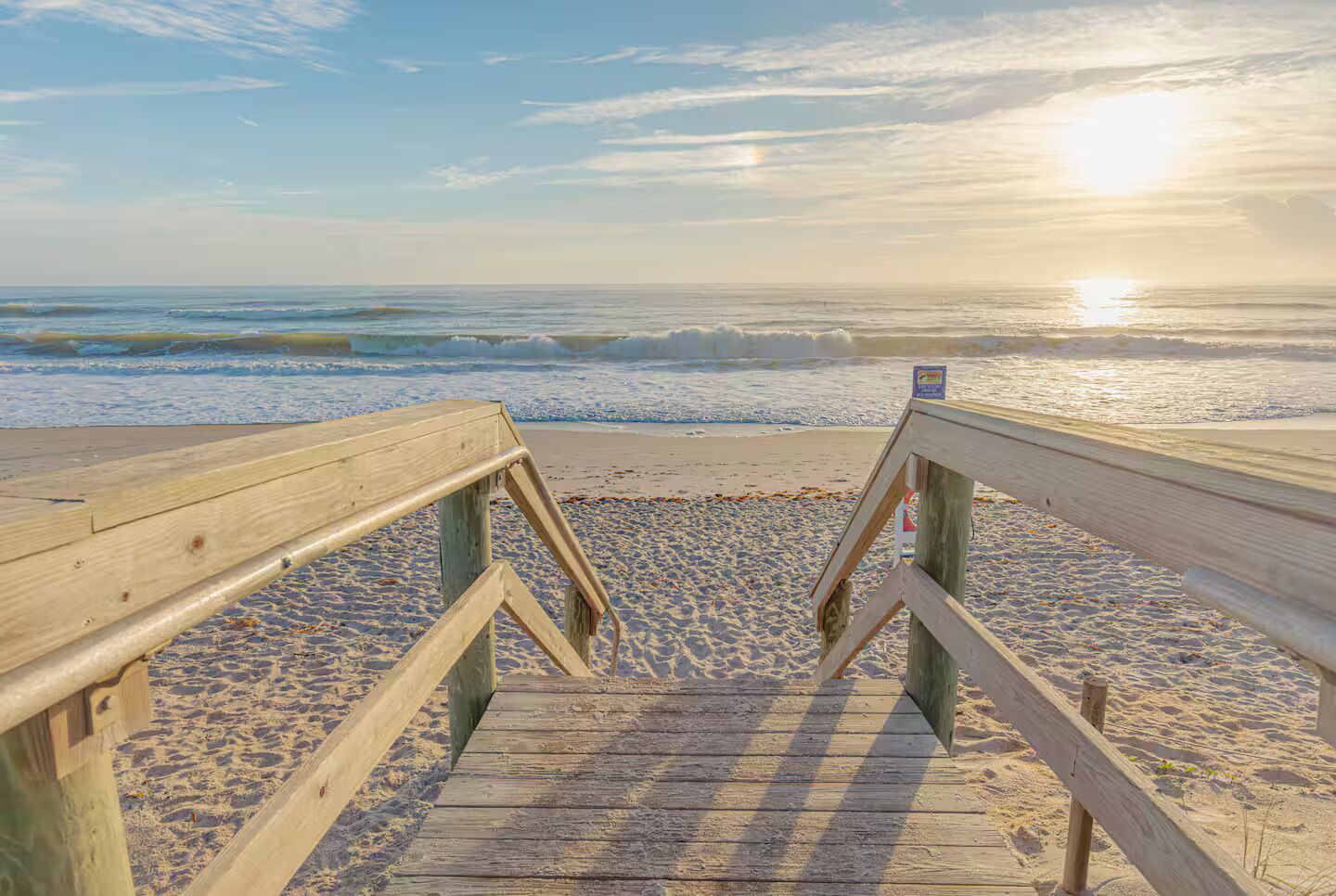 Ein hölzerner Steg führt zu einem Strand, wo Wellen brechen und die Sonne im Hintergrund untergeht.