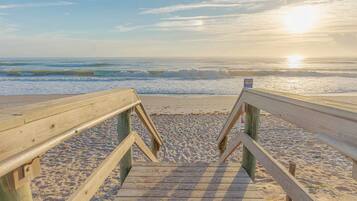 Ein hölzerner Steg führt zu einem Strand, wo Wellen brechen und die Sonne im Hintergrund untergeht.