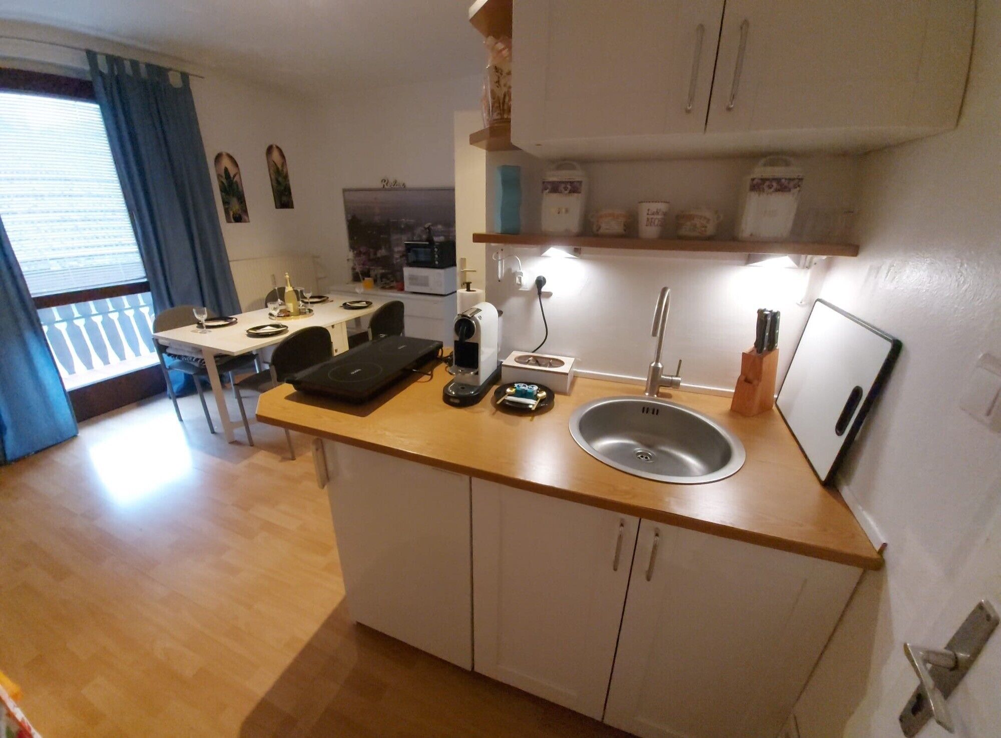 A compact kitchen with a sink, countertop, and cabinets. There is a dining table with chairs and a window with curtains.