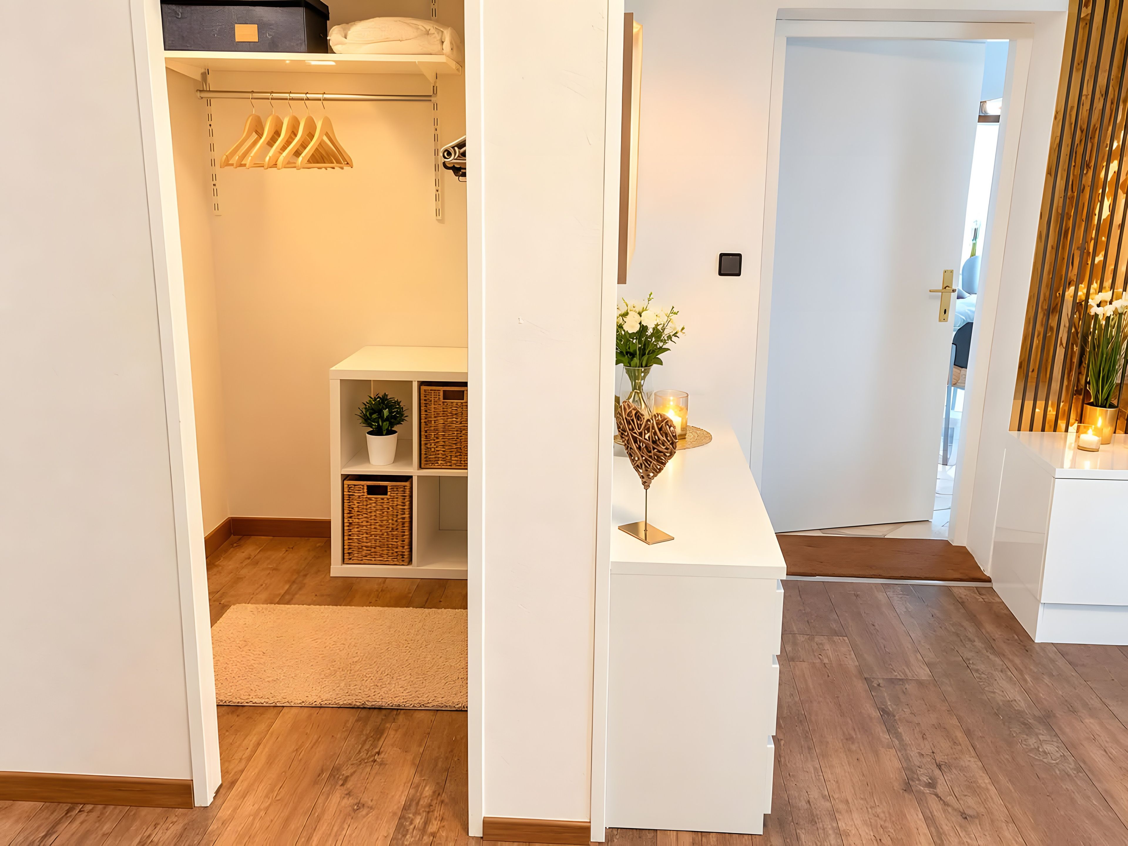A hallway with a white console table, a potted plant, and a candle. There is a wooden floor and a door leading to another room.