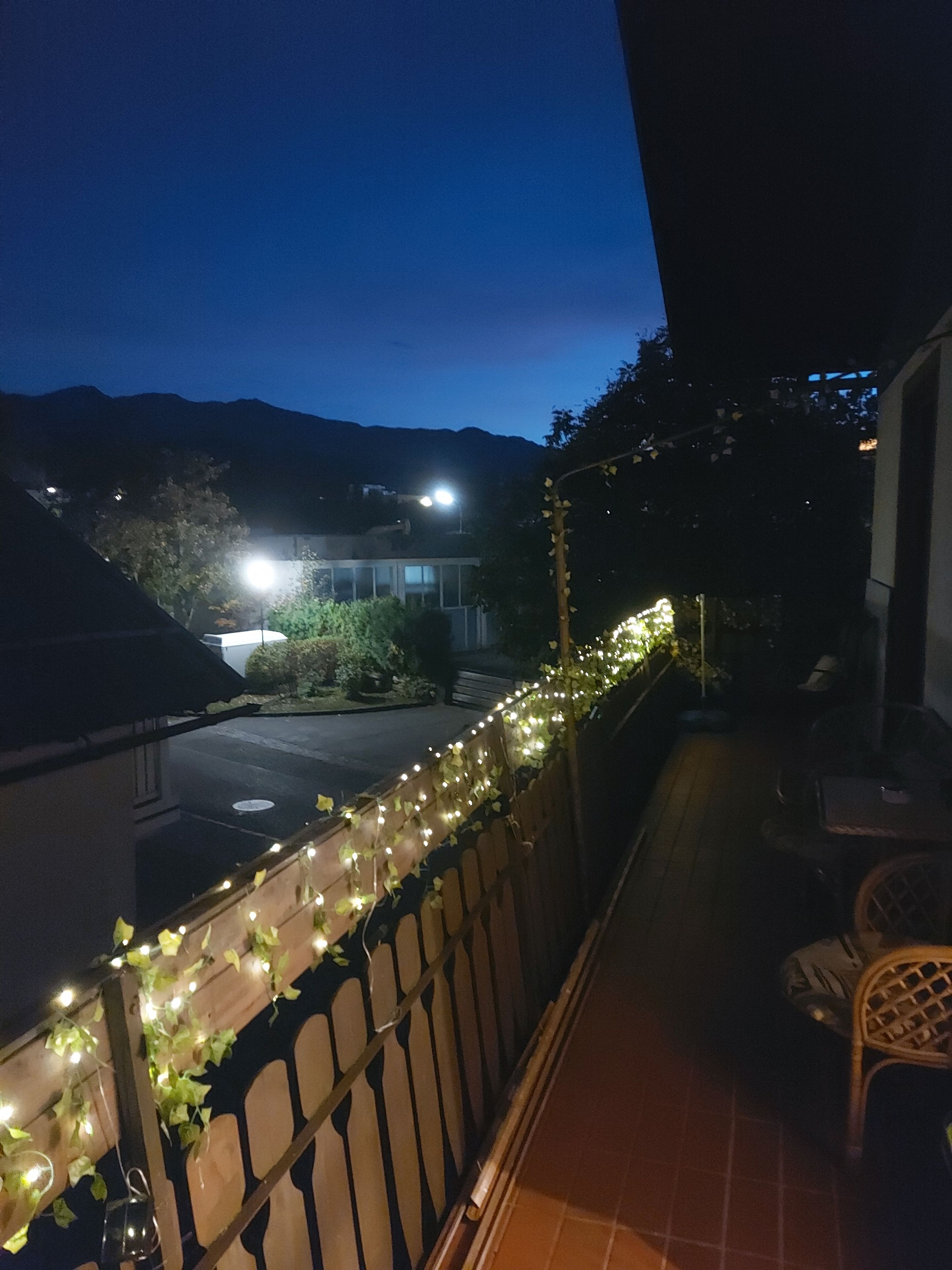 A balcony with a view of a street and mountains, decorated with string lights and ivy.