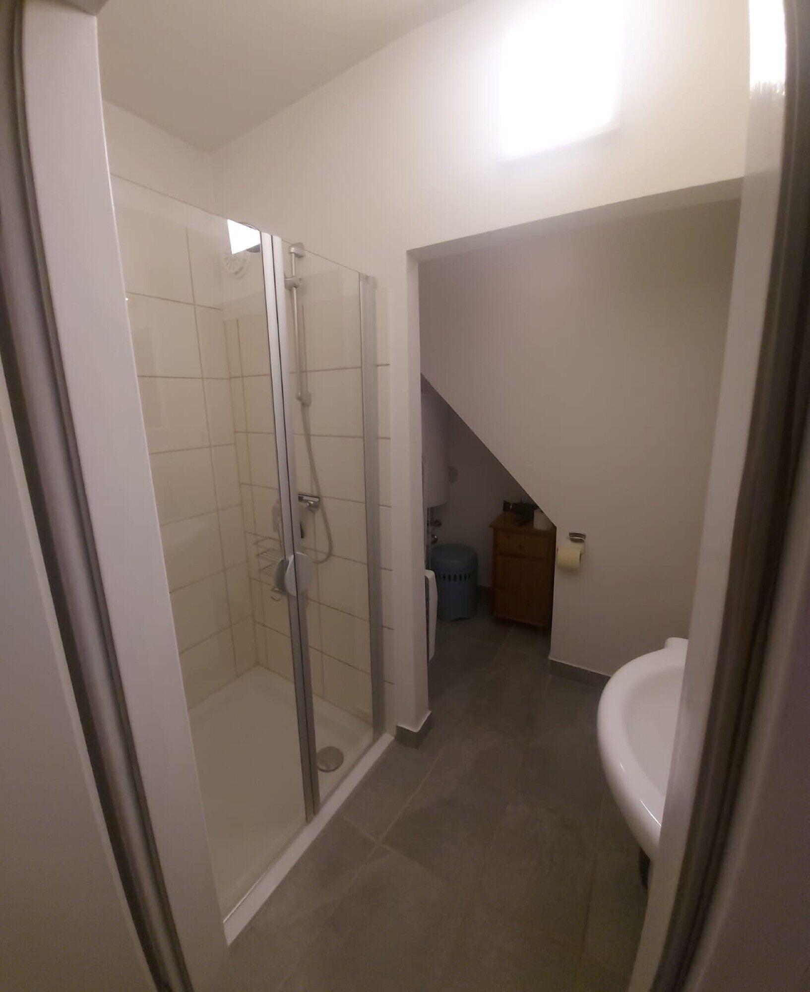 Een badkamer met een glazen douchecabine, een wit wastafeltje en een houten kastje.