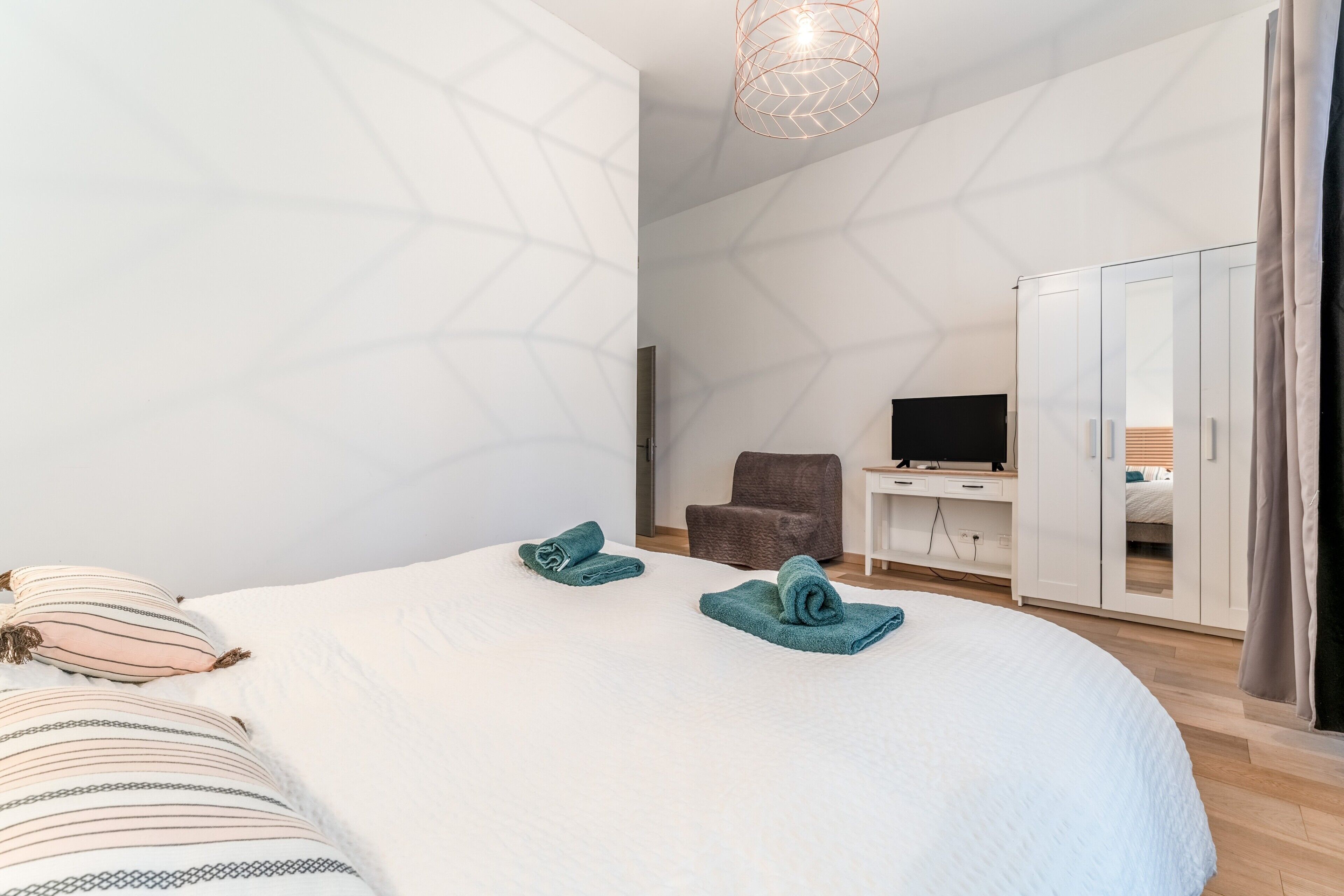 A modern hotel room with a large bed, a desk with a TV, a wardrobe, and a geometric-patterned wall.