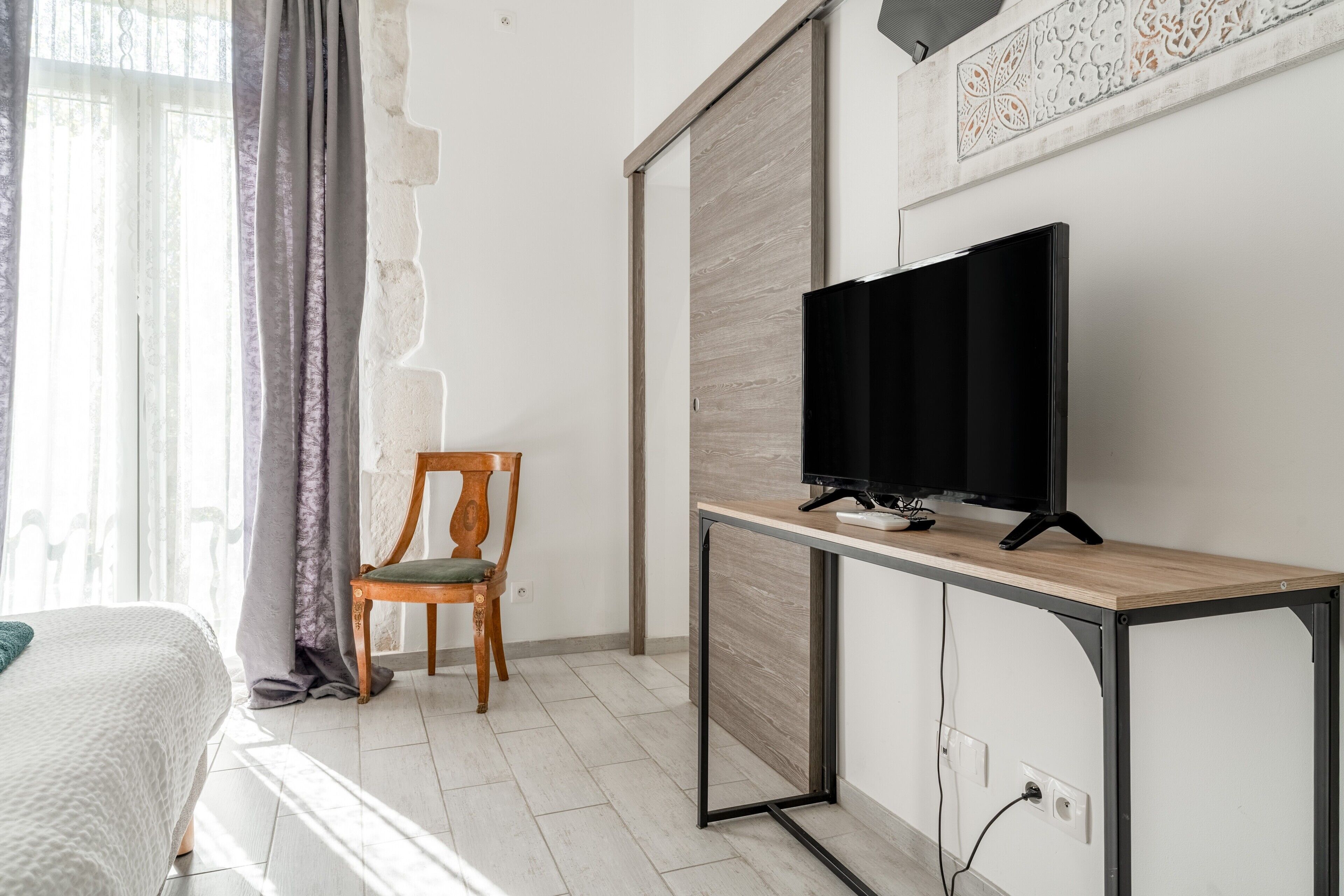Una stanza con un letto, una sedia in legno, una TV su una scrivania in legno e una porta scorrevole.