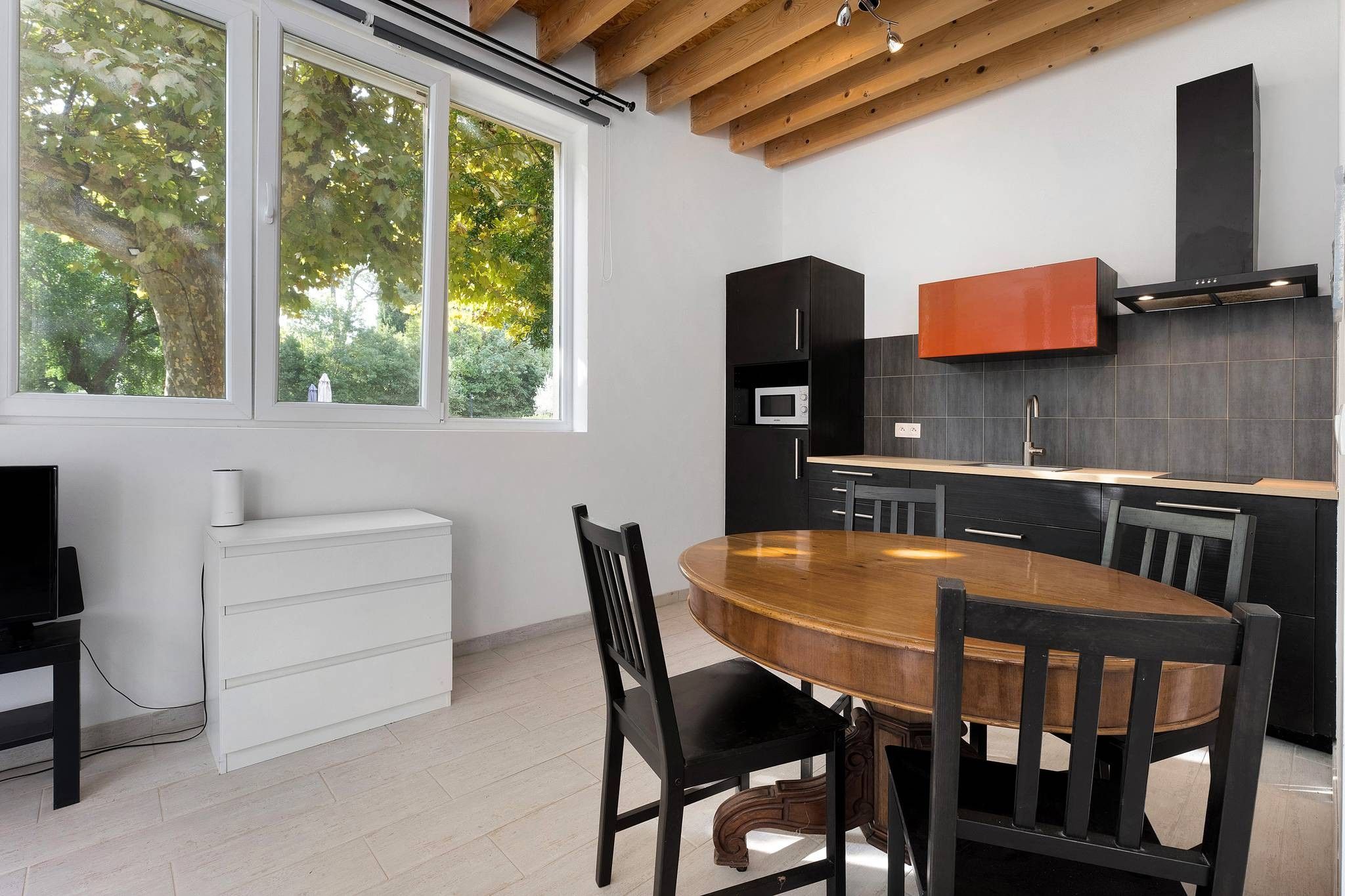 A modern kitchen with a wooden dining table and chairs, a built-in oven, and a large window with a view of trees.