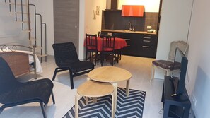 A modern living room with a sofa, two round tables, a black chair, a dining area with a red tablecloth, and a kitchen in the background. - Charming Studio Le Mas de Kitty, Shared Garden and Pool (Saint-Rémy-de-Provence)