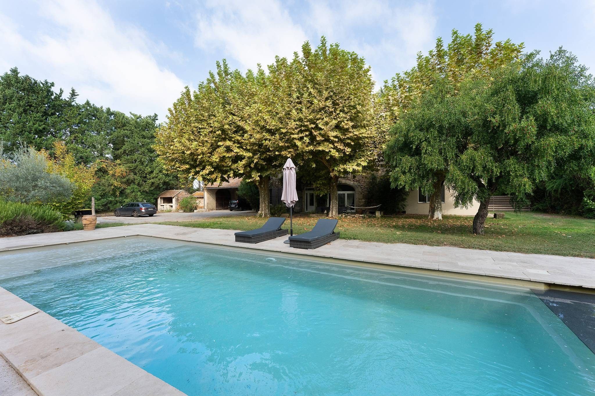 A swimming pool with lounge chairs and a covered umbrella, surrounded by trees and a building.