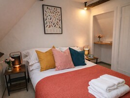 A neatly made bed with a red blanket and multiple pillows, a framed bird illustration on the wall, a bedside lamp, and a small nightstand with a vase of flowers.