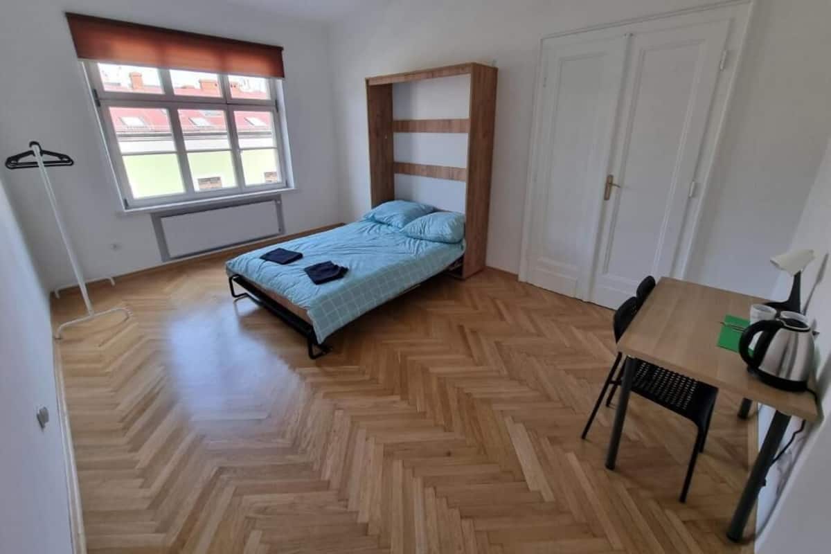 A bedroom with a bed, a table, and a chair. There is a window with blinds and a radiator.