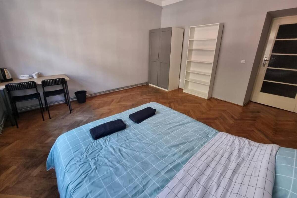 A bedroom with a bed, a desk with chairs, a wardrobe, and a shelving unit.