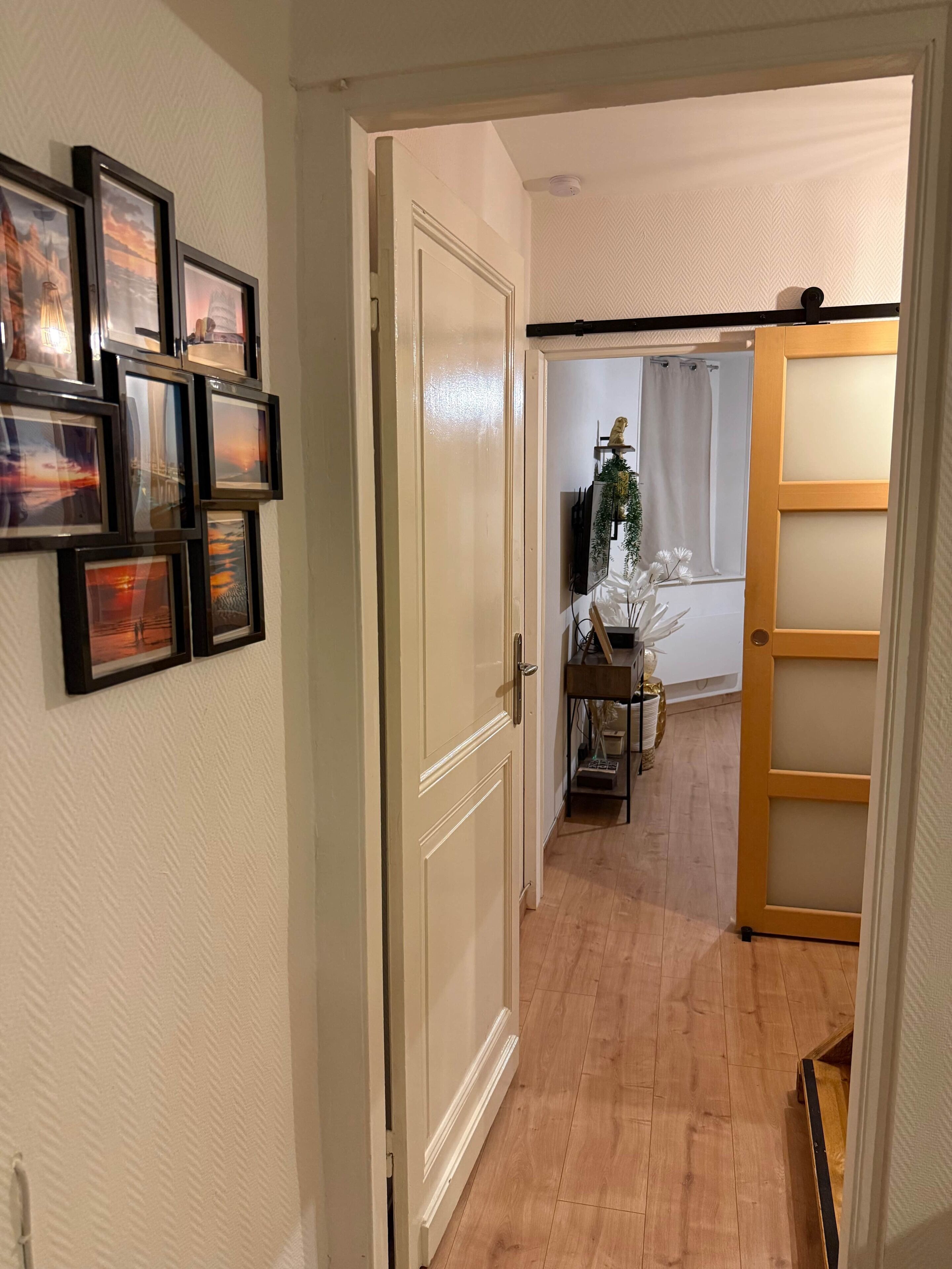 Un couloir avec des tableaux encadrés accrochés au mur, une porte en bois et une vue sur une pièce où l’on aperçoit une table et des rideaux.